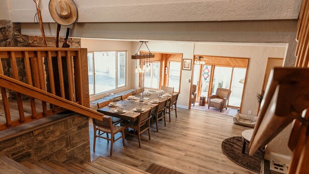 A spacious dining area with a long wooden table set for six, flanked by wooden chairs. A staircase with a wooden railing leads to an upper level.