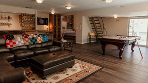 A living room with a pool table, a leather sofa, a dartboard, and a staircase leading to a balcony.
