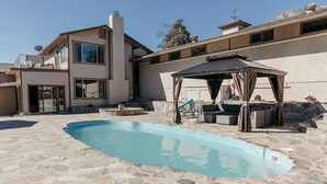 A swimming pool with stone tiles, a covered seating area, and a modern house with a balcony.