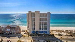 Een hoog hotel met meerdere verdiepingen, uitzicht op het strand en een parkeerplaats ervoor.
