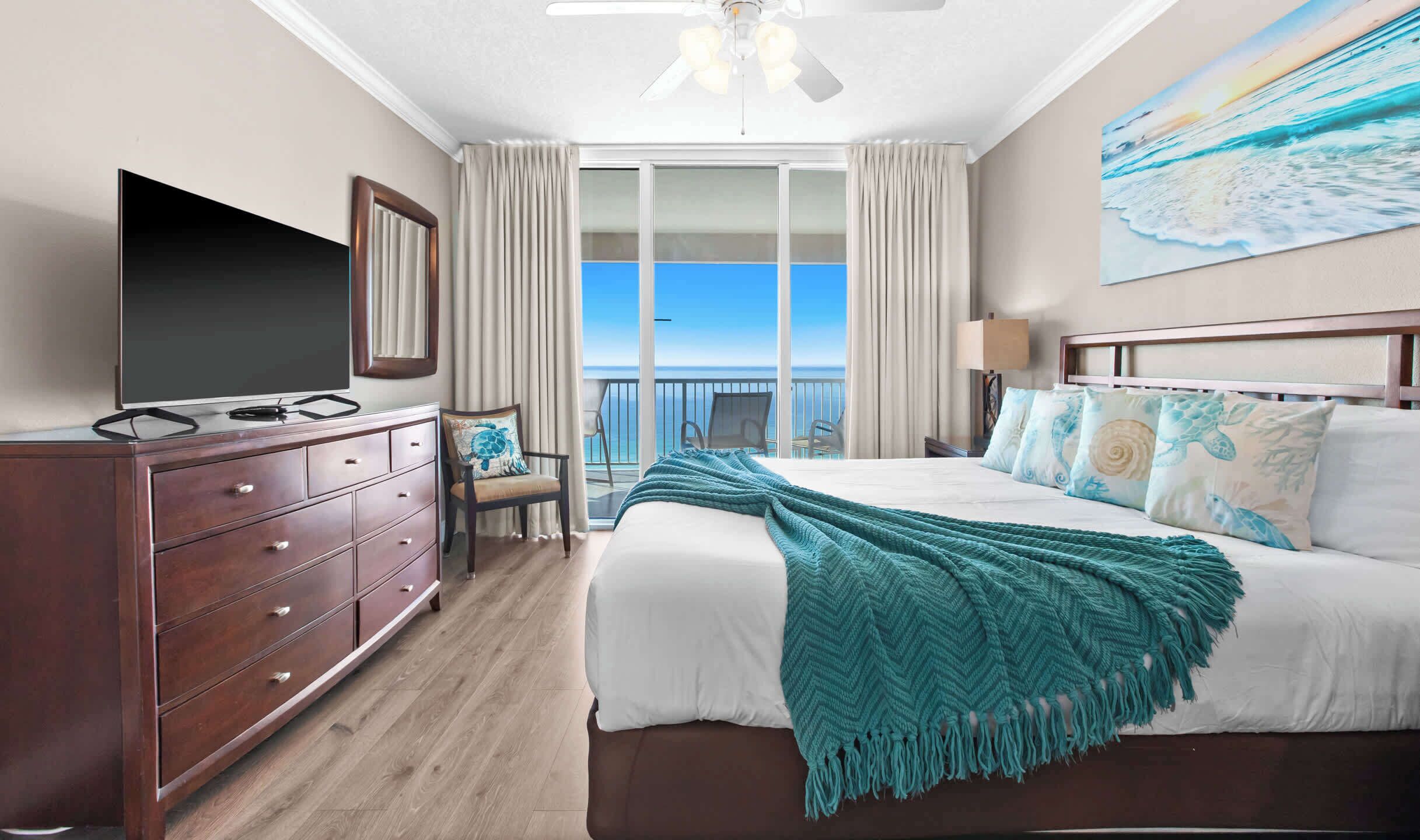 A hotel room with a large bed, a TV, a dresser, and a balcony with a view of the ocean.