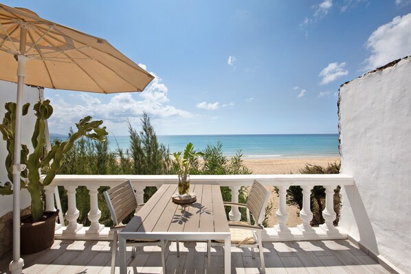 A balcony with a table, chairs, a potted plant, and an umbrella overlooking a beach.