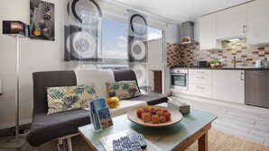 En moderne stue med en sofa, et sofabord med en dekorativ tallerken, og et køkkenområde med hvide skabe og et flisebeklædt bagpanel.