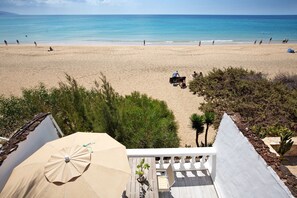 A beach view from a balcony with a wooden deck, outdoor seating, and a beach umbrella.