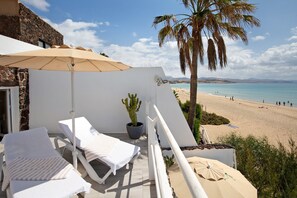 A balcony with white lounge chairs and a beige umbrella, overlooking a beach with clear blue water and a palm tree.