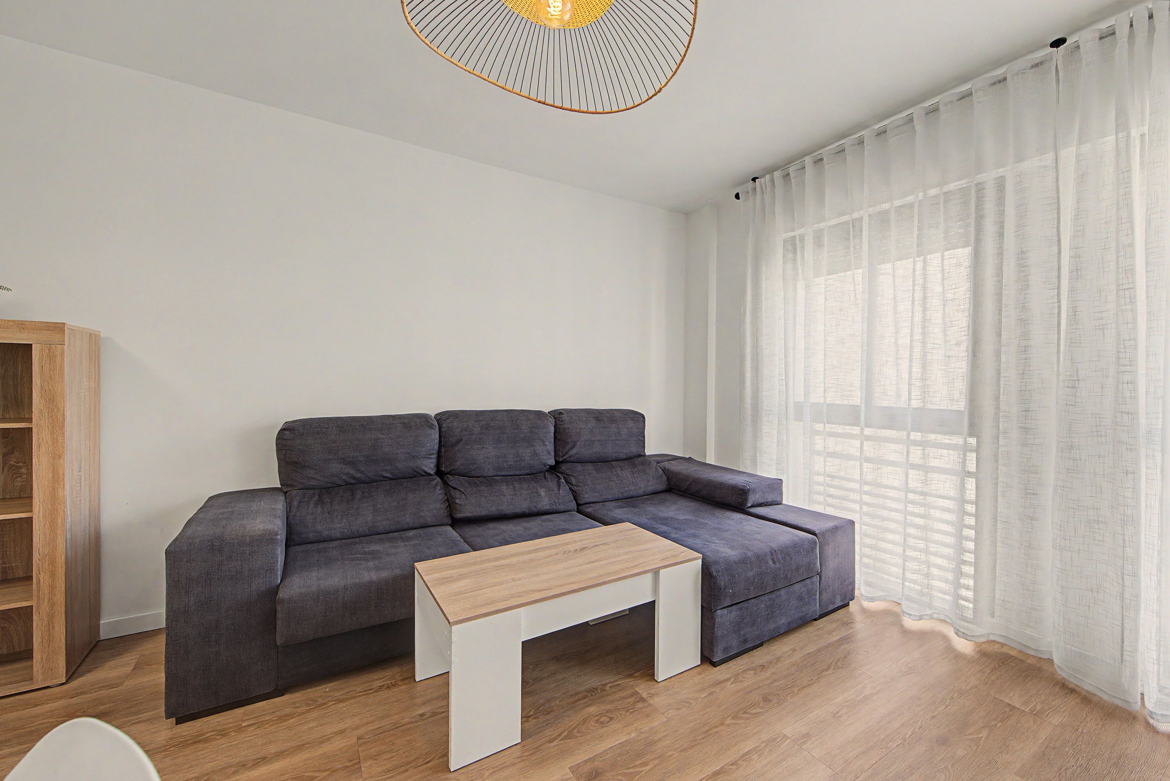 Una sala de estar moderna con un sofá seccional gris, una mesa de centro de madera y un gran ventanal con cortinas translúcidas.