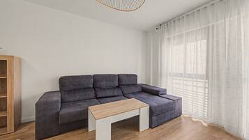 Una sala de estar moderna con un sofá seccional gris, una mesa de centro de madera y un gran ventanal con cortinas translúcidas.