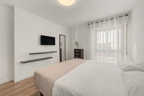 Un dormitorio con cama, televisor montado en la pared, cómoda y ventana con cortinas.
