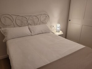 Un lit bien fait, avec du linge de lit blanc, une tête de lit blanche ornée de motifs circulaires, une table de chevet avec une lampe et une porte fermée.