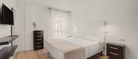 Un dormitorio con cama, mesitas de noche, televisor y ventana con cortinas.