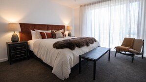 Hotellihuone, jossa on suuri sänky, kaksi yöpöytää valaisimineen, penkki, tuoli ja suuri ikkuna, jossa on ohuet verhot.