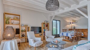 A spacious living room with a television, bookshelves, a dining area with a table set for four, and a large window. - 4-Star Villa Arcachon – Heated Pool, Garden & Petanque, Sleeps 8 (La Teste-de-Buch)