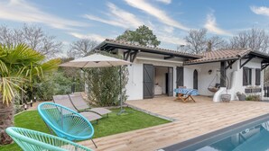 A poolside area with a wooden deck, two blue chairs, a hanging chair, and a covered seating area. - 4-Star Villa Arcachon – Heated Pool, Garden & Petanque, Sleeps 8 (La Teste-de-Buch)
