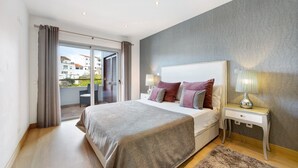 Une chambre avec un grand lit, une table de chevet avec une lampe, une vue sur le balcon extérieur et un mur gris à la texture intéressante.