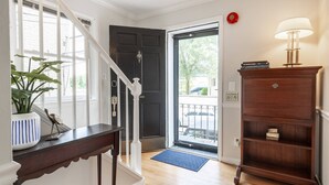 A hallway with a wooden staircase, a dark door, a blue mat at the entrance, a wooden cabinet, and a lamp. - Steps to USNA Gate 3, On-Site Parking & Private Patio at Historic Gatehouse (Annapolis)