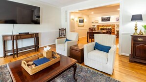 A living room with a TV, two white armchairs, a wooden coffee table, and a wicker side table. - Steps to USNA Gate 3, On-Site Parking & Private Patio at Historic Gatehouse (Annapolis)