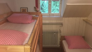A small, cozy room with two wooden beds, each with a checkered pillow. A radiator is visible in the center, and a window with a view of greenery is on the wall. - Bergwaldhütte  (Bühl)