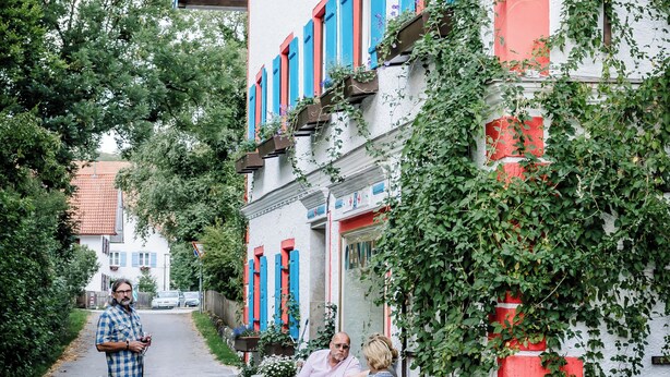A narrow street with a white building featuring red and blue shutters, potted plants, and a wooden cross.