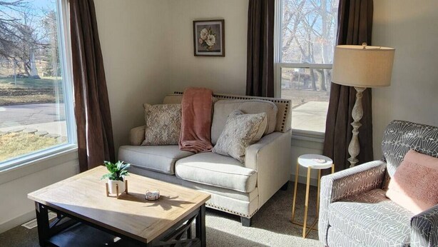 A living room with a sofa, a coffee table, a lamp, and a chair. There is a window with curtains, a picture on the wall, and a view of the outside. - Cozy American Style Cottage (Sioux City)