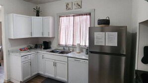 A compact kitchen with white cabinets, a stainless steel refrigerator, a window with curtains, and a small plant on the counter. - Cozy American Style Cottage (Sioux City)