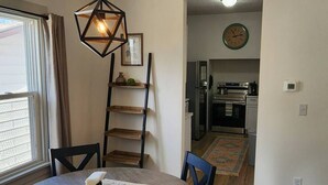 A dining area with a round table, chairs, a wooden ladder shelf, a hanging light fixture, and a kitchen in the background. - Cozy American Style Cottage (Sioux City)