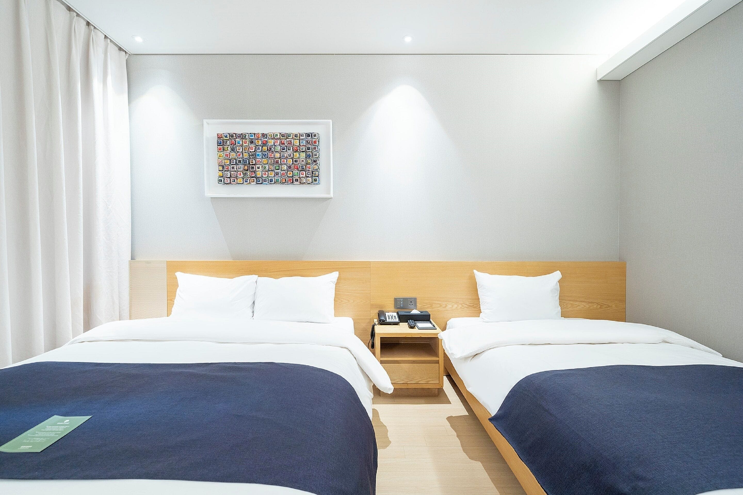 A hotel room with two beds, a wooden headboard, a nightstand with a phone and a book, and a framed picture on the wall.