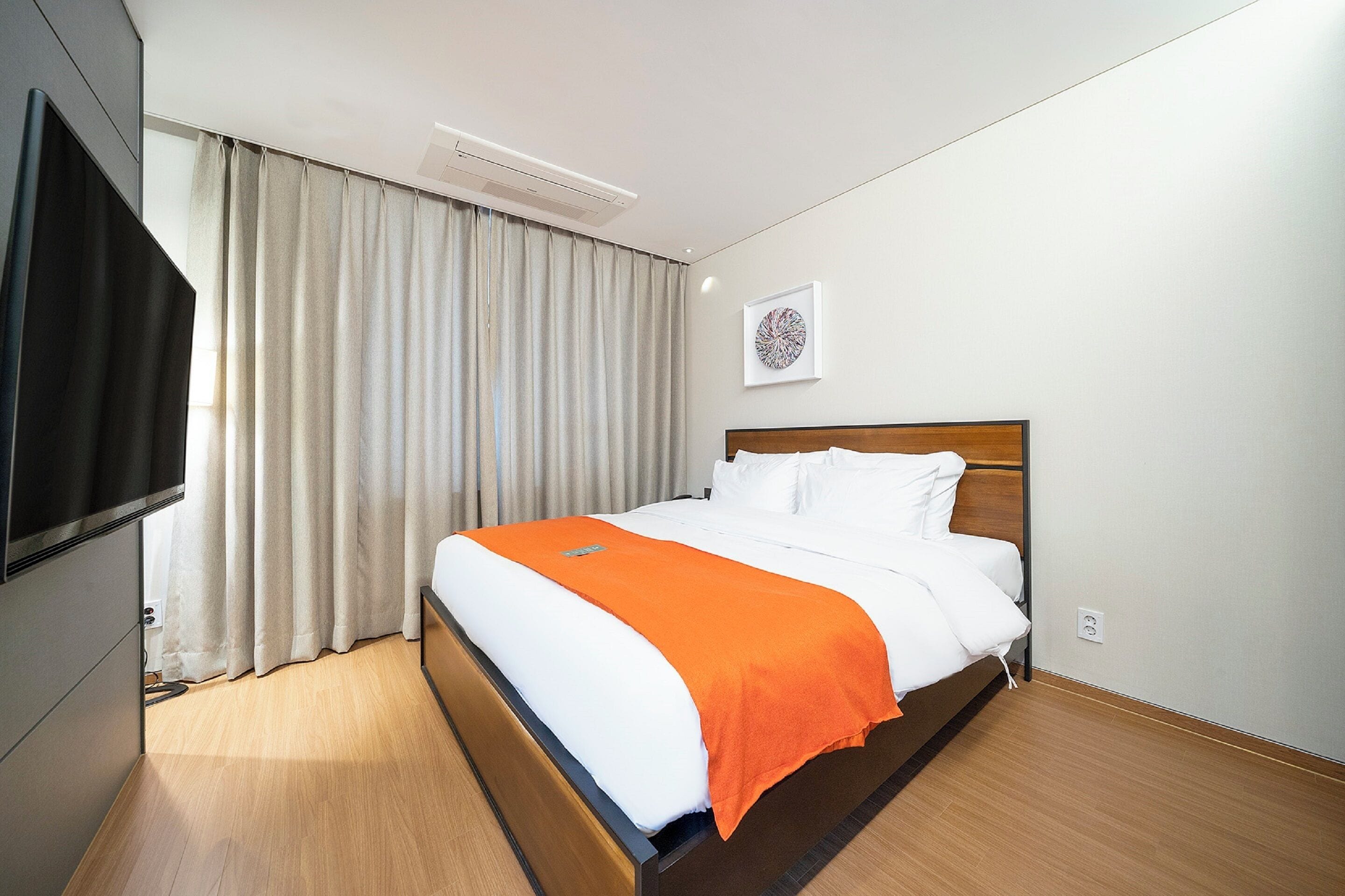 A hotel room with a bed, a TV, a window with curtains, and a wall-mounted artwork.