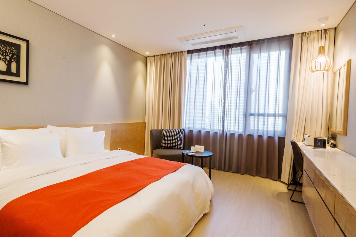 A hotel room with a large bed, a chair, a small table, and a window with curtains.