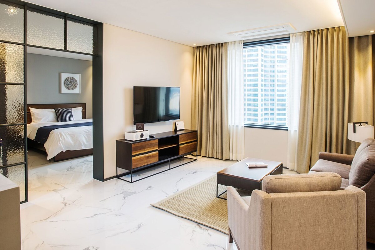 A hotel room with a bed, a TV, a sofa, a coffee table, and a large window with curtains.