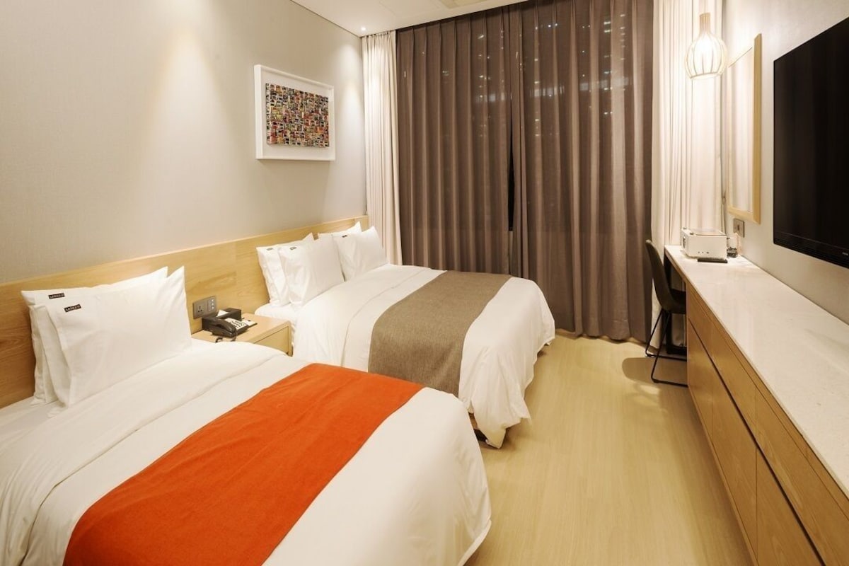 A hotel room with two beds, a desk, a TV, and a large window with curtains.