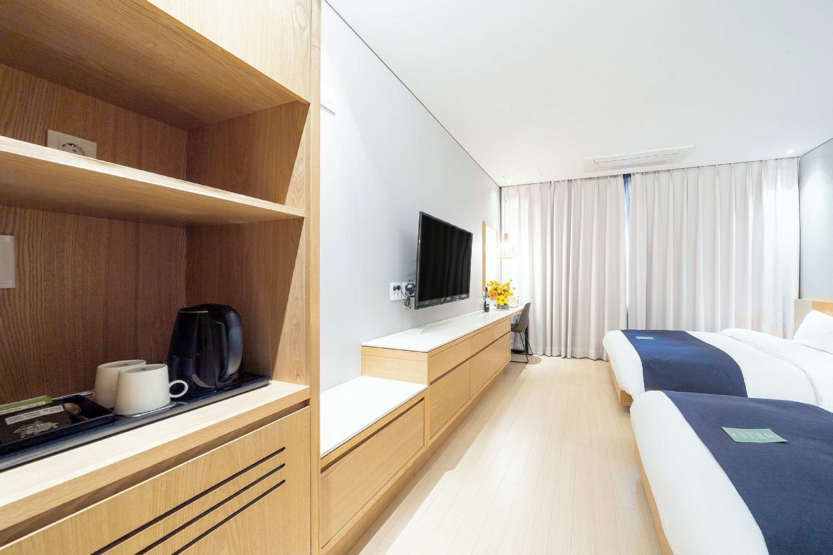 Habitación de hotel con armario de madera, televisor de pantalla plana, mesita de noche con cafetera y cama con manta azul.