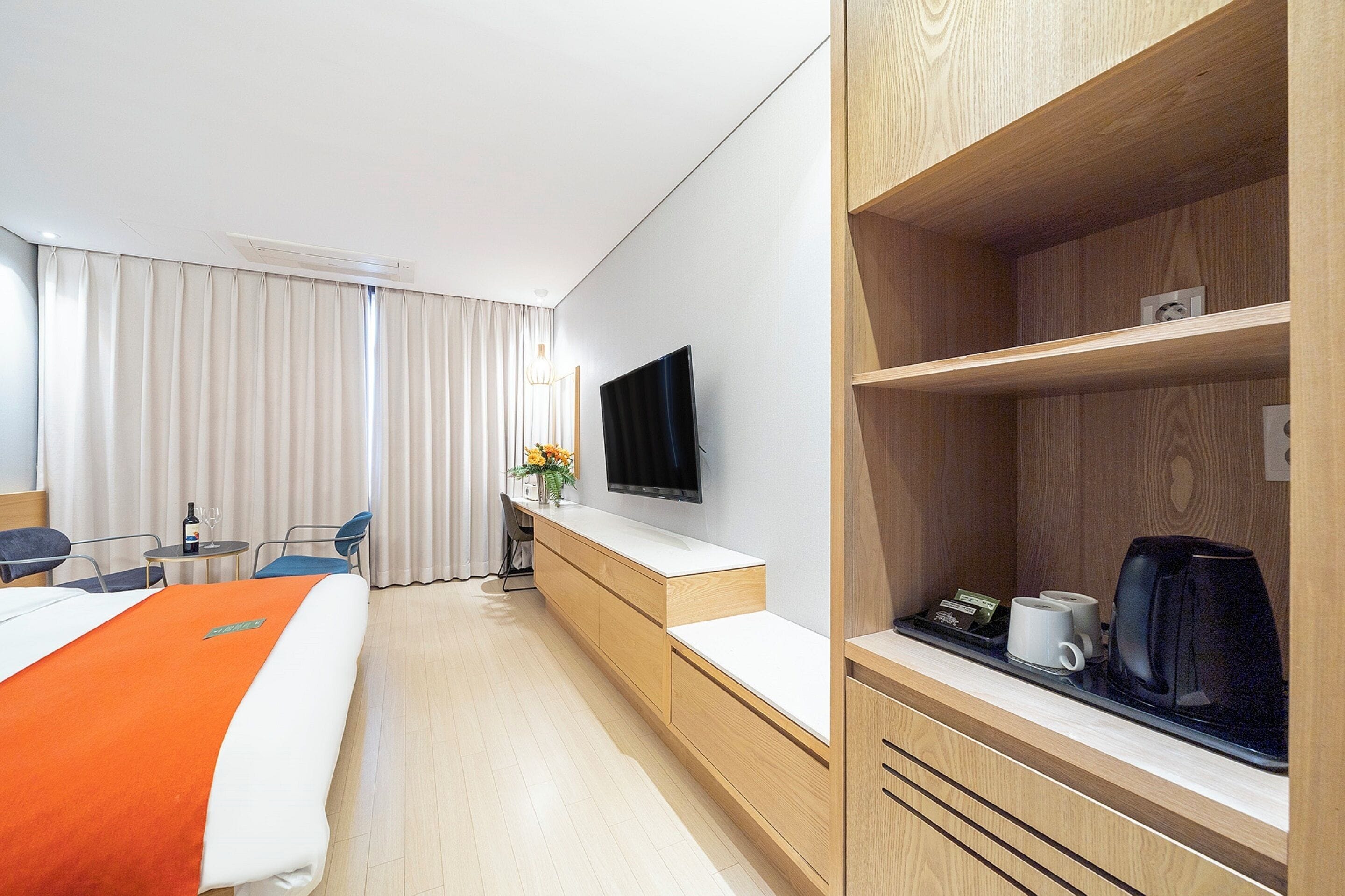 A hotel room with a bed, a desk with a flat-screen TV, a coffee maker, and a shelf with a kettle.