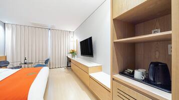A hotel room with a bed, a desk with a flat-screen TV, a coffee maker, and a shelf with a kettle.