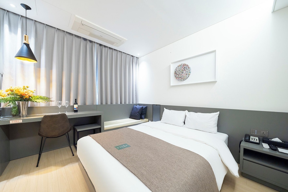A modern hotel room with a bed, desk, chair, and a bedside table with a phone.