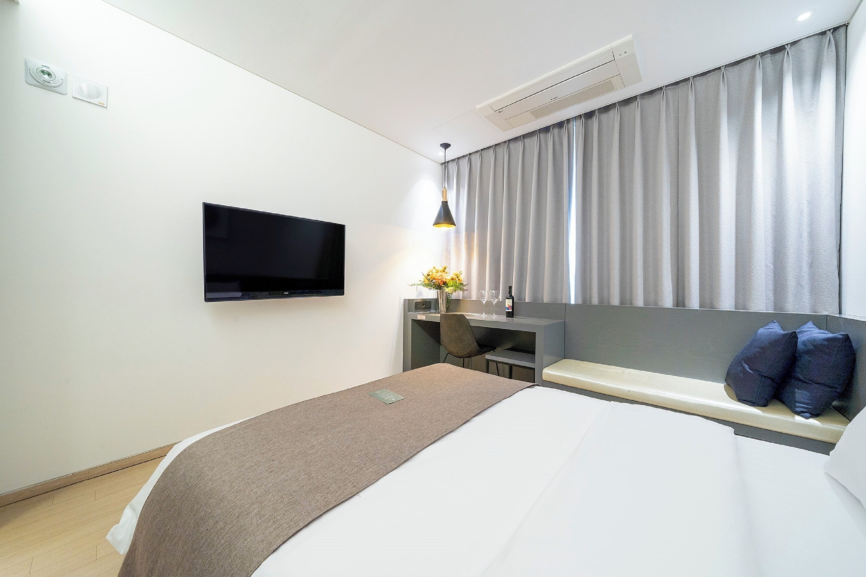 Une chambre d’hôtel moderne équipée d’un lit, d’un téléviseur fixé au mur, d’un bureau avec une chaise et d’une grande fenêtre avec des rideaux.