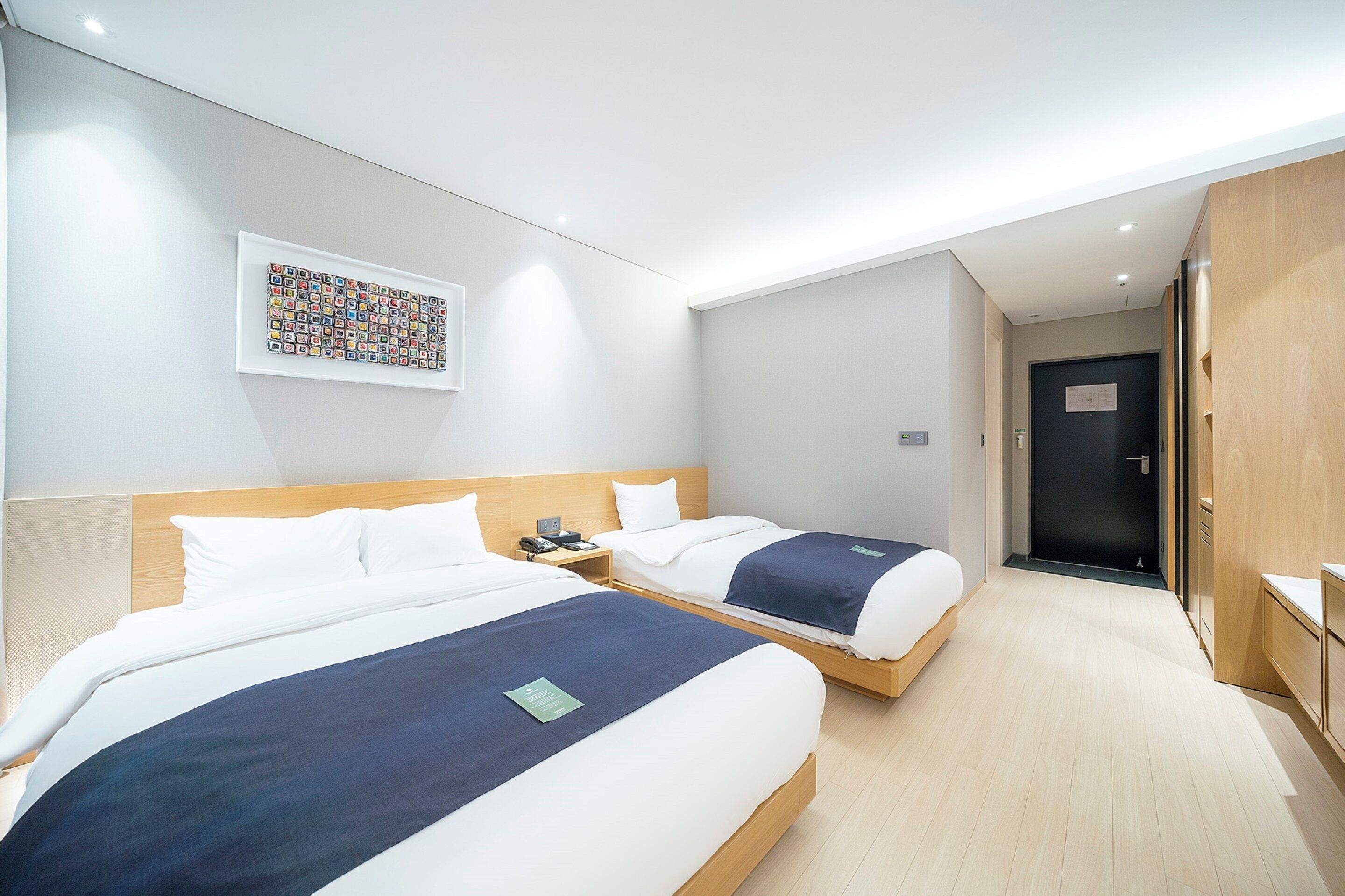 A hotel room with two beds, a wooden headboard, a wall-mounted artwork, and a door leading to a hallway.