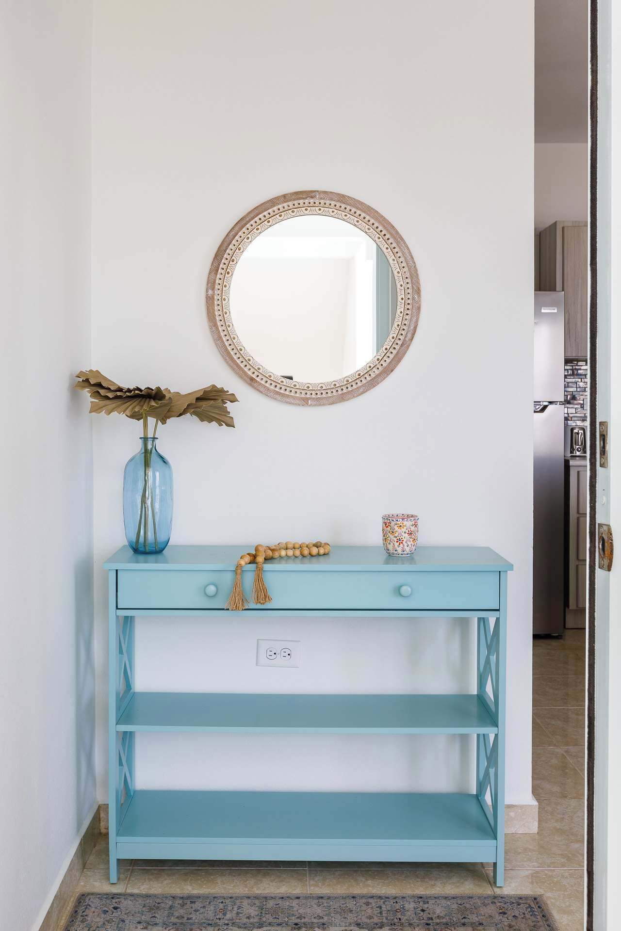 A light blue console table with a round mirror above it, a vase with dried branches, a wooden prayer bead, and a patterned mug.