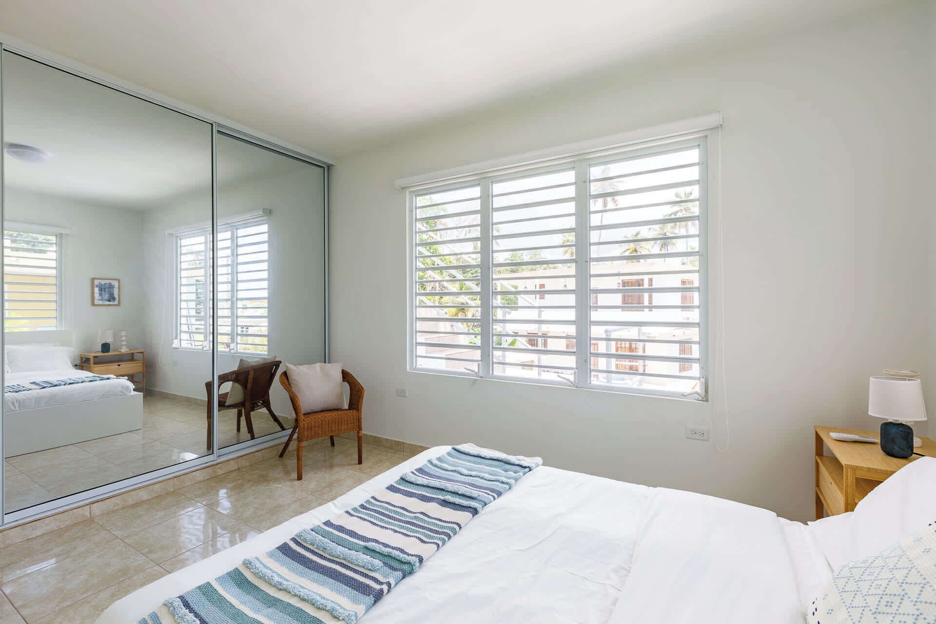A bedroom with a bed, a chair, a nightstand, a lamp, and a window with blinds.