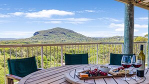 Una mesa de madera con una tabla de quesos, uvas y copas de vino en un balcón con vistas a una montaña y un bosque.