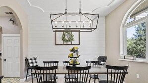 A dining area with a rectangular table, six black chairs, a chandelier, a framed plant, and a window with a view of trees. - Spacious-Gourmet Kitchen-Luxe King Bed-Ping Pong-Arcade (Sonora)