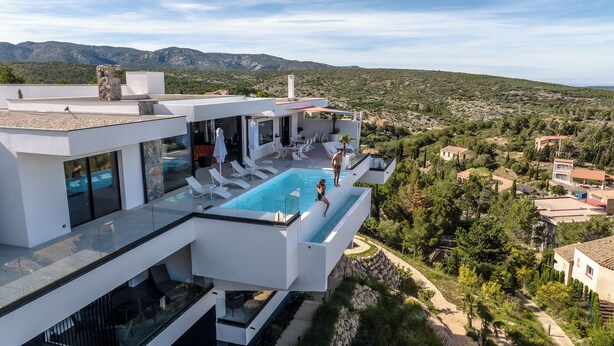 Une villa moderne dotée d’une piscine à débordement sur le toit, d’une terrasse fermée par des baies vitrées et offrant une vue panoramique sur un paysage montagneux.