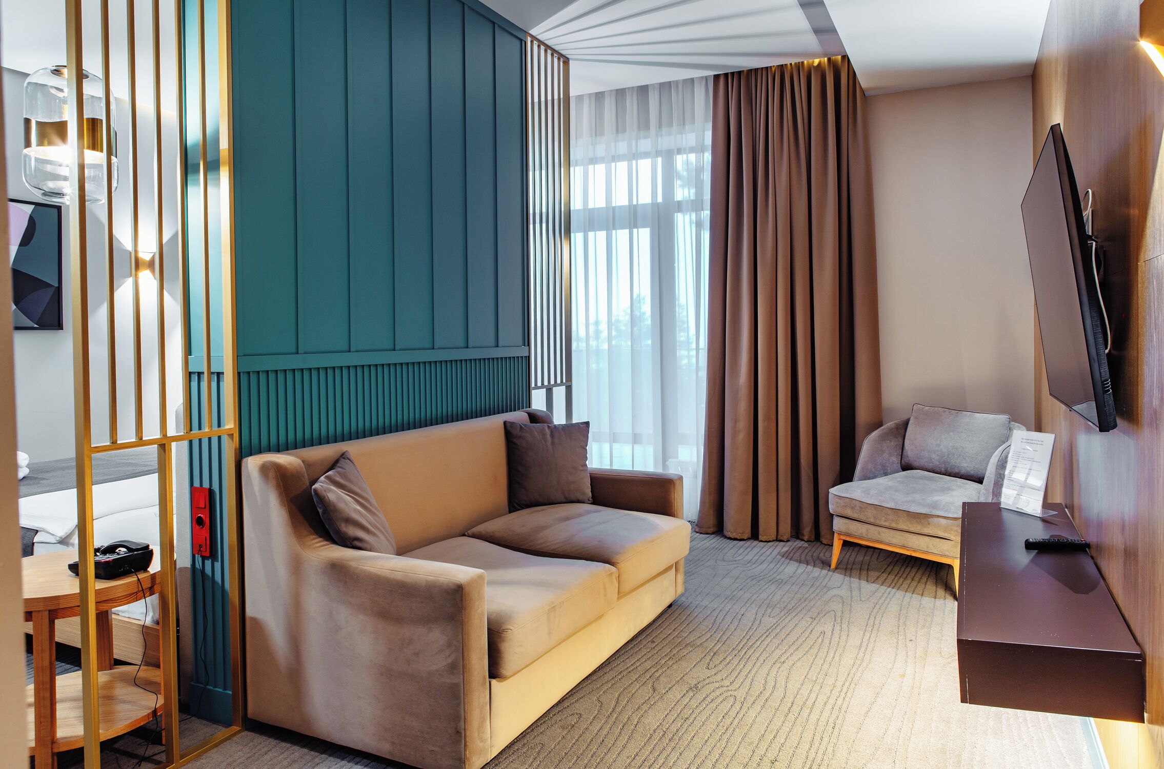 Camera d'hotel moderna con divano, una poltroncina, televisore a schermo piatto, scrivania in legno e un'ampia finestra con tende.