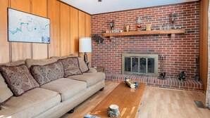 Ein Wohnzimmer mit Kamin aus Backstein, Holzwandverkleidung, einer Couch mit gemusterten Kissen und einer Karte an der Wand.