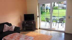 A living room with a sofa, a television on a stand, a table with a floral tablecloth, and a sliding glass door leading to an outdoor patio area. - Holiday home in Thale in the Harz Mountains (Thale ot Allrode)