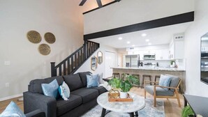 A modern living room with a sofa, coffee table, and chairs, a staircase, and a kitchen in the background.