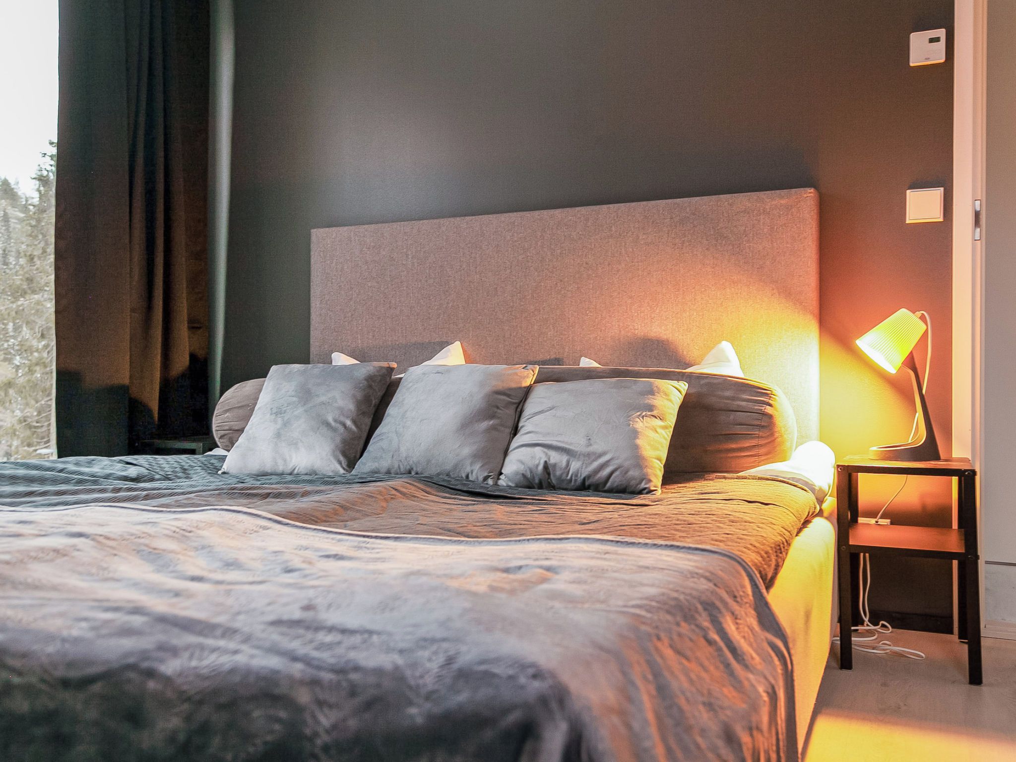 A modern bedroom with a large bed, multiple pillows, a bedside table with a lamp, and a view of the outdoors.