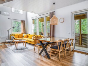 A bright living room with a yellow sofa, wooden dining table, and chairs. There are large windows with curtains, a hanging light fixture, and a clock on the wall. - Saukonpesä a by Interhome (Kuusamo)