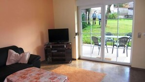 A living room with a sofa, a coffee table, a TV on a stand, and a sliding glass door leading to an outdoor patio area with chairs. - Cozy apartment in Thale. (Thale ot Allrode)