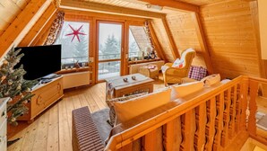 A cozy attic room with a wooden interior, a Christmas tree, a sofa, a chair, and a television.