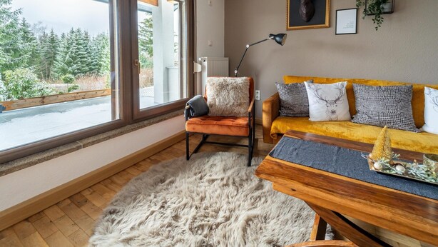 Ein gemütliches Wohnzimmer mit großem Fenster, einem flauschigen Fellteppich, einem Holztisch und einer Couch mit gemusterten Kissen.